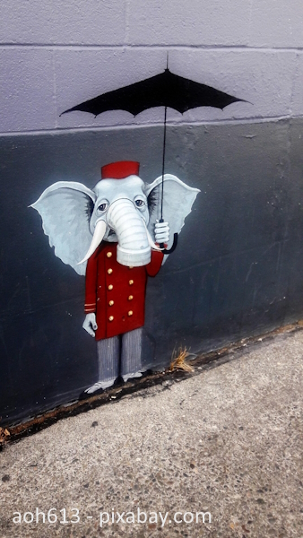 Auf eine graue Wand ist ein Elefant gemalt. Er steht aufrecht wie ein Mensch und trägt eine rote Jacke mit Goldknöpfen. Aus der Wand ragt ein gebogenes Rohr, das zum Rüssel wird.