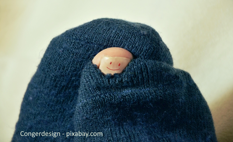 Zwei Füße in dunkelblauen Socken. In einem ist im Bereich des großen Zehs ein Loch. Der Zeh schaut heraus. Auf den Nagel hat jemand ein lächelndes Gesicht gemalt.