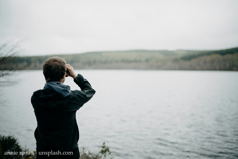 Eine Person steht am See. Mit einem Fernglas sieht sie in die Ferne. Vielleicht ist sie auf der Suche nach einem neuen Anfang.